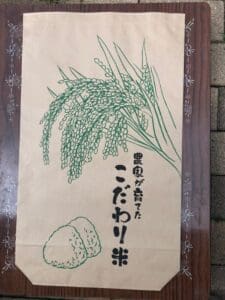 稲穂とおにぎりの緑色のイラストと「ごろへい米」「コシヒカリ」という日本語の文字が描かれた茶色の紙袋が木の表面に置かれている。
