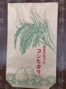 緑色の稲穂と底におにぎり2個が描かれた茶色の紙袋。赤い日本語で「ごろへい米」と書いてある。袋は暗い模様の木の上に置かれています。