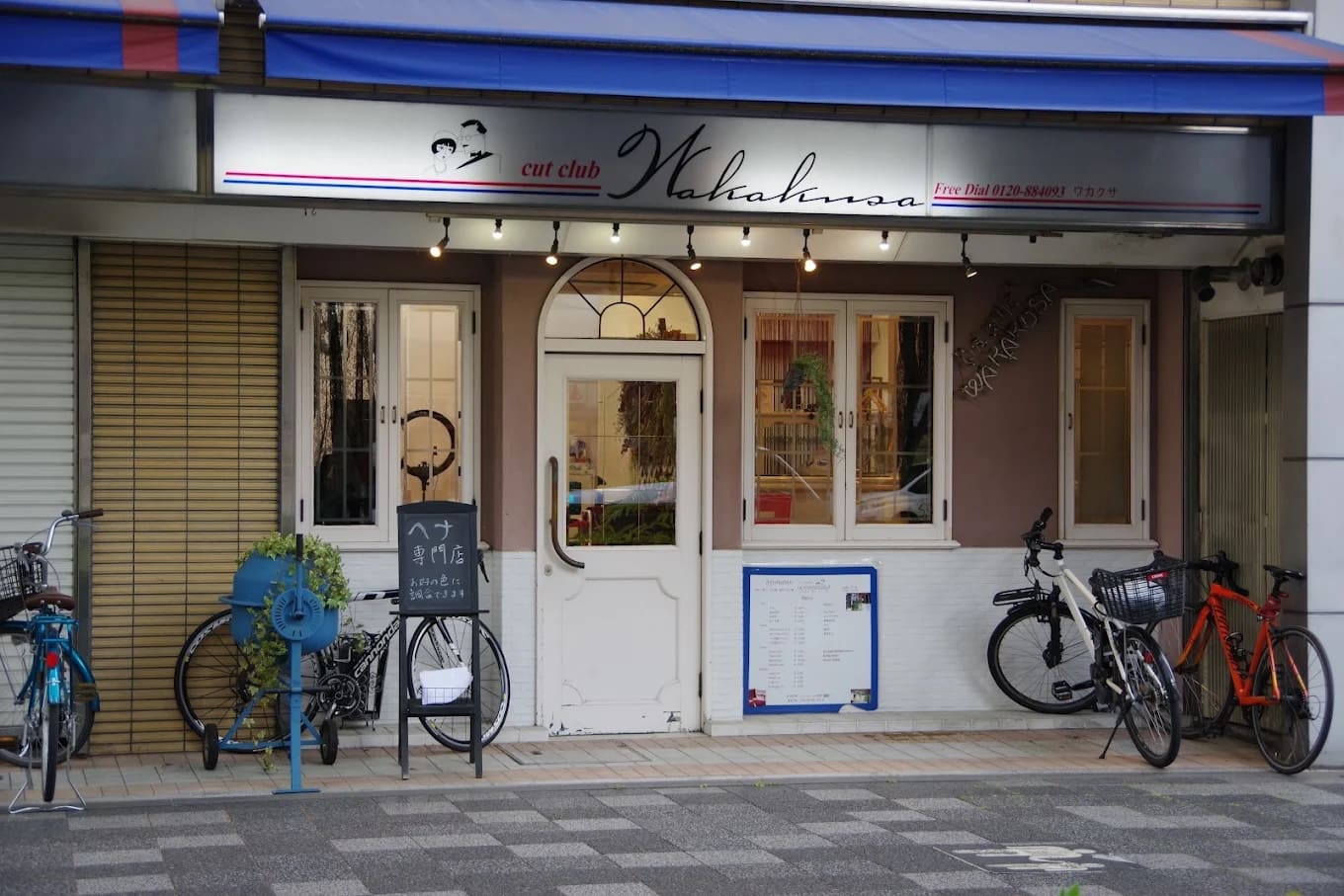 Der Laden hat ein Schild mit der Aufschrift "Cut Club Wakana" auf einer weißen Doppeltür und bietet Abkürzungen und Henna-Services an. Zwischen den Fenstern und dem Fahrradparkplatz befinden sich blaue Fahrradständer, Zimmerpflanzen sowie Speisekarten und Schilder in der Nähe des Eingangs.