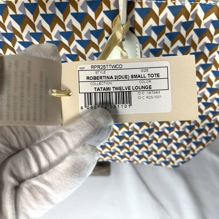 Die behandschuhte Hand hält das Produktetikett "ROBERTINA 2 (DUE) SMALL TOTE" auf dem Stoff mit Tatami-Muster mit einem geometrischen Muster in Blau, Braun und Weiß, dem charakteristischen Stil der kleinen Tote-Kollektion von ROBERTA PIERI.