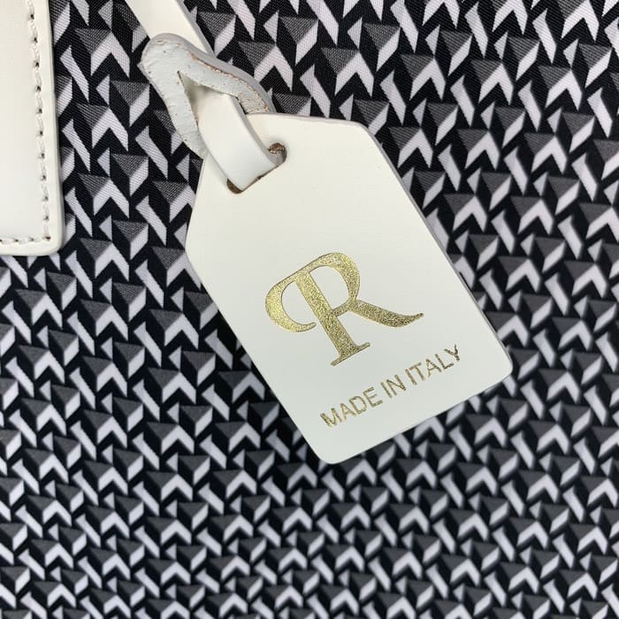 Ein weißes ROBERTA PIERI-Etikett aus Leder mit den Buchstaben "R" in Gold und den Worten "MADE IN ITALY" hängt vor dem Hintergrund eines schwarz-weißen geometrischen Stoffes.