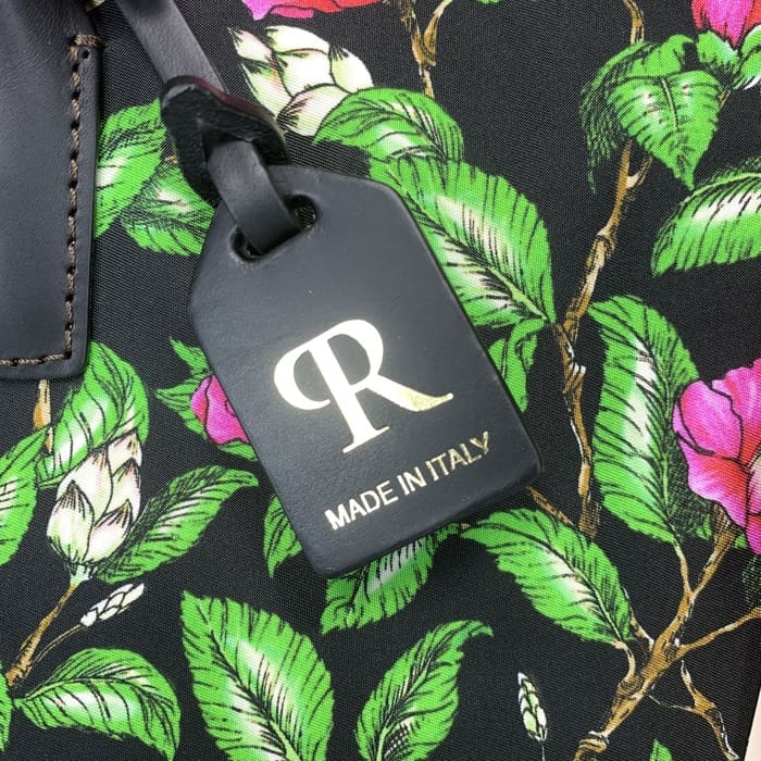 Der schwarze ROBERTA PIERI Kofferanhänger mit dem goldenen "R" und dem italienischen Text hängt an einem floralen Stoff mit grünen Blättern und rosa Blumen und ergänzt die Mini-Reisetasche mit zeitloser italienischer Eleganz.