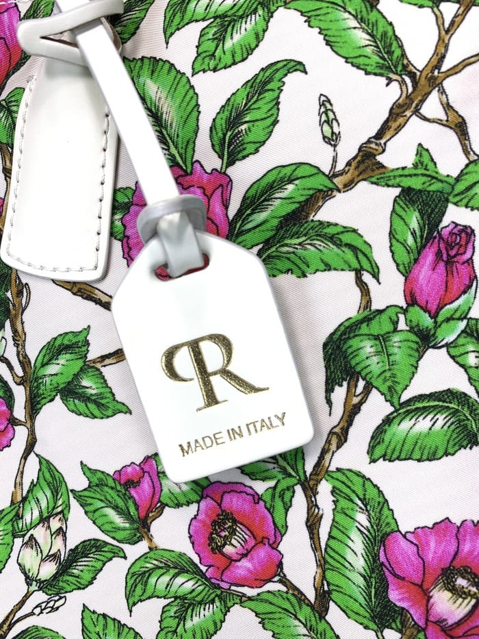 Nahaufnahme der ROBERTA PIERI Mini-Reisetasche. Stiele mit grünen Blättern und rosa Blüten; Das weiße Etikett an der 2-Wege-Tasche ist mit einem goldenen "R"-Logo und dem Schriftzug "MADE IN ITALY" versehen.