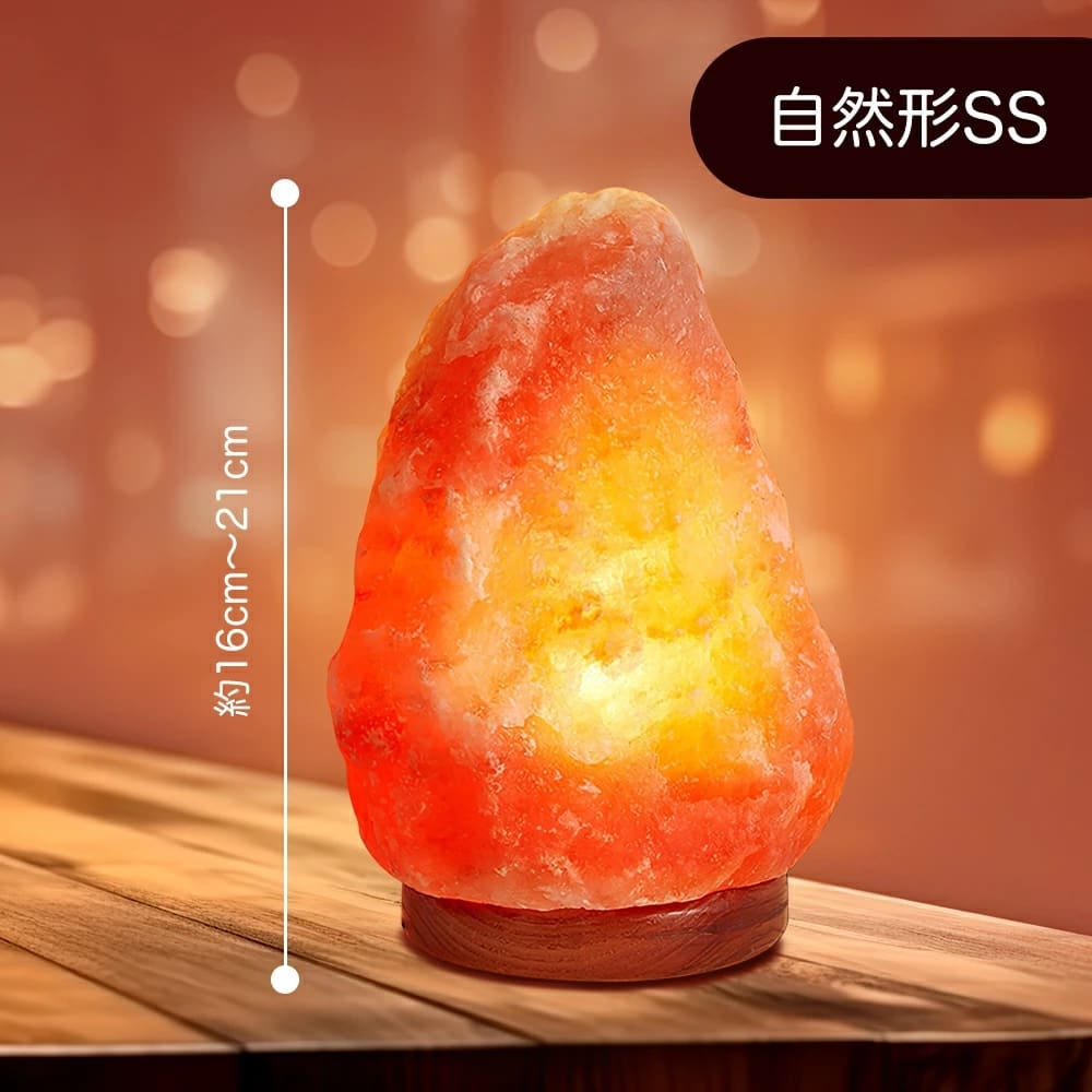 オレンジ色に輝く天然岩塩ランプ【サイズ：SS】— ほっと落ち着く、優しい間接照明が、木製の台座の上に載せられ、テーブルに飾られています。 この天然岩塩ランプは周囲を柔らかく照らし、穏やかな間接照明の役割を果たしています。白い文字で高さが約16〜21cmと記されています。右上隅には漢字が書かれた黒いラベルが貼られています。.